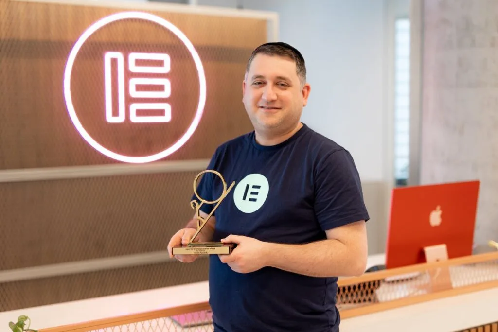 Elementor co-founder holding an award trophy in front of a neon Elementor logo in the company office