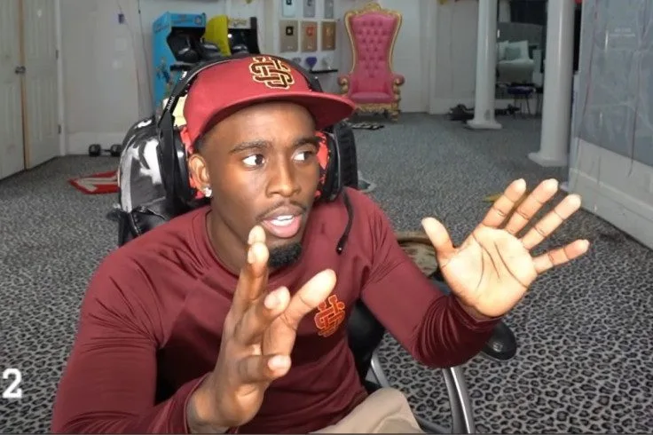 Kai Cenat in a red cap and headset gesturing with both hands while streaming from his room